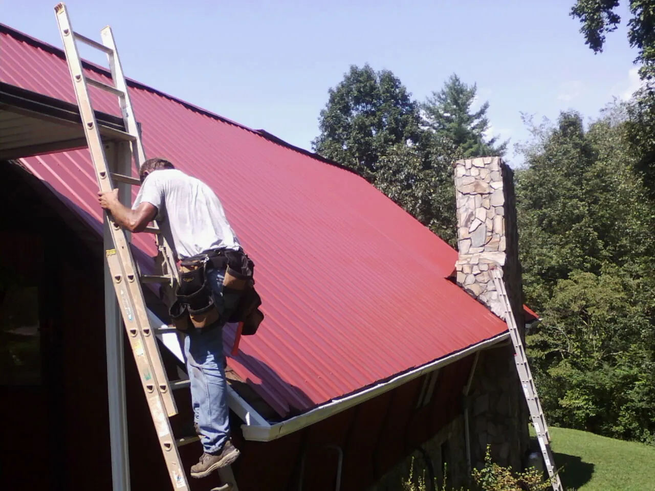 Roofer on a ladder beside a red metal roof and stone chimney — Soffit & Fascia Repair by Roofing Brothers in Neffs